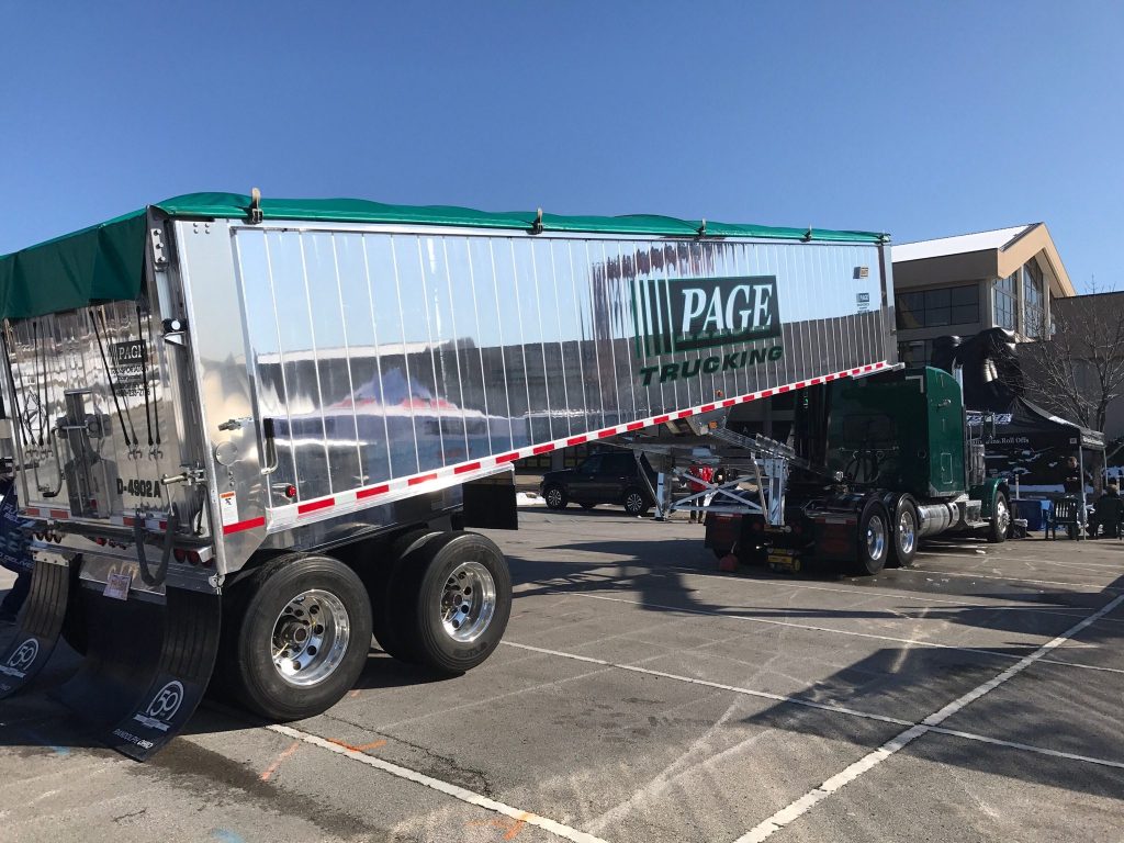 A shiny silver semi-trailer truck with green accents parked outdoors, featuring the logo 'PAGE TRUCKING' on its side, with a clear blue sky in the background.